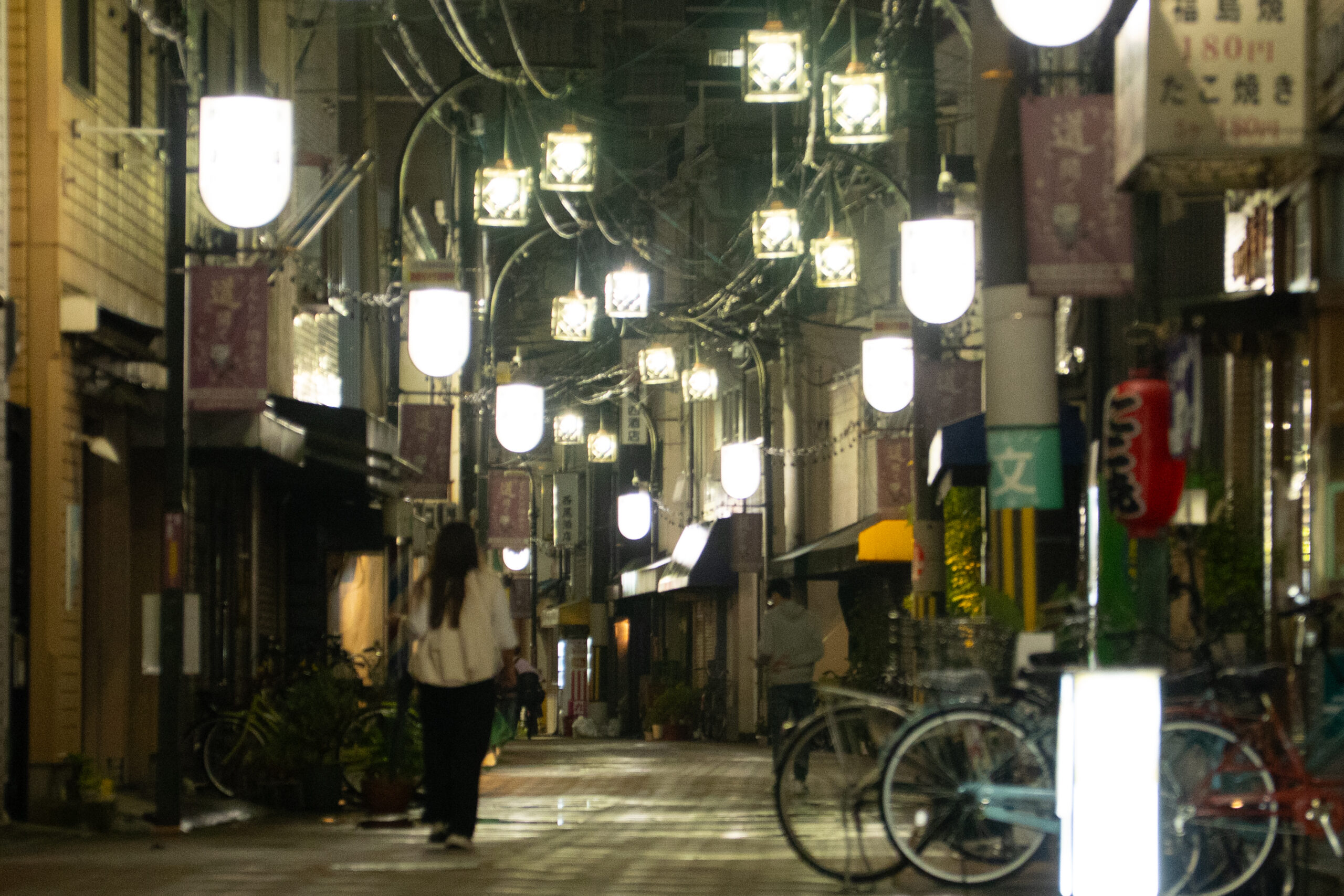 A covered shopping street at night, with hanging lights, bicycles and people walking